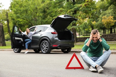 Upset man sitting near warning triangle and broken car on road