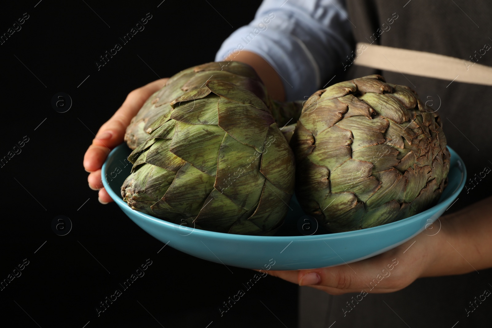 Photo of Woman holding bowl with fresh raw artichokes on black background, closeup