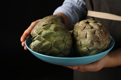 Photo of Woman holding bowl with fresh raw artichokes on black background, closeup