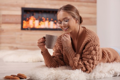 Beautiful young woman with cup of hot drink on rug at home. Winter atmosphere