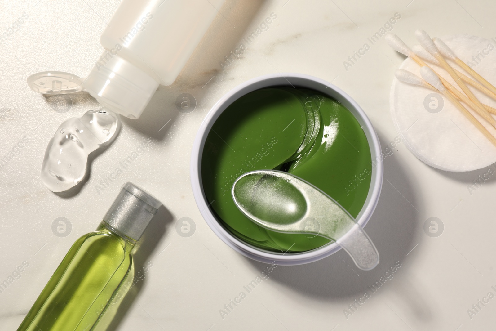 Photo of Jar of under eye patches with spoon and cosmetic products on white marble table, flat lay