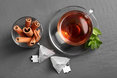 Tea bags, cup of hot drink, mint and cinnamon sticks on grey wooden table, flat lay