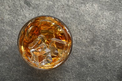 Photo of Whiskey and ice cubes in glass on grey textured table, top view. Space for text