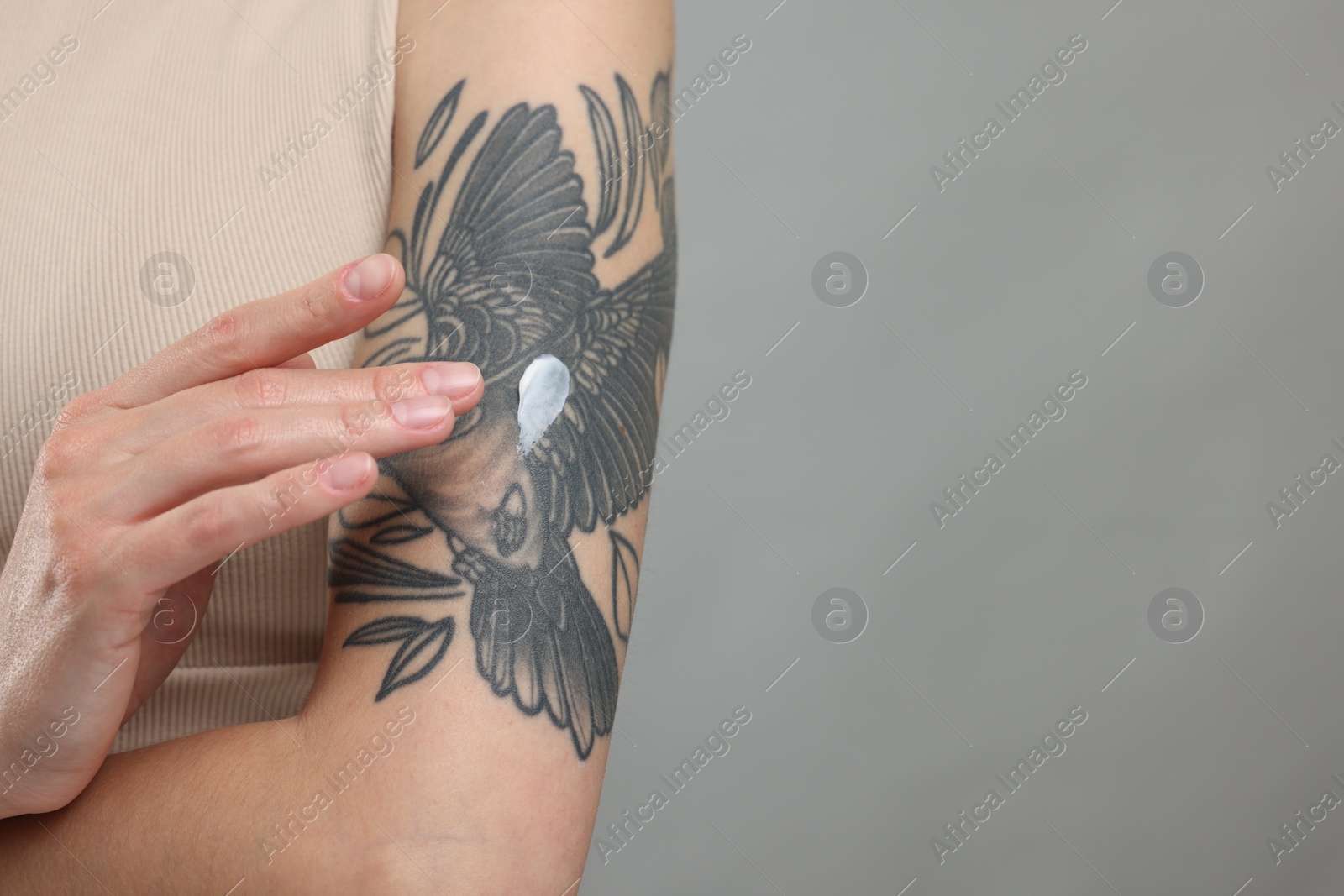 Photo of Tattooed woman applying cream onto her arm on gray background, closeup. Space for text