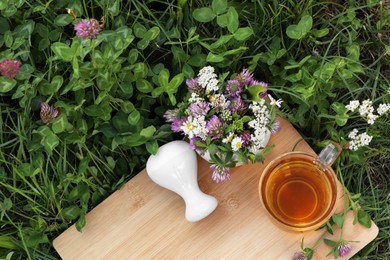 Photo of Cup of aromatic herbal tea, pestle and mortar with different wildflowers on green grass outdoors, flat lay. Space for text