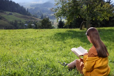 Photo of Young woman reading book on green meadow in mountains, back view