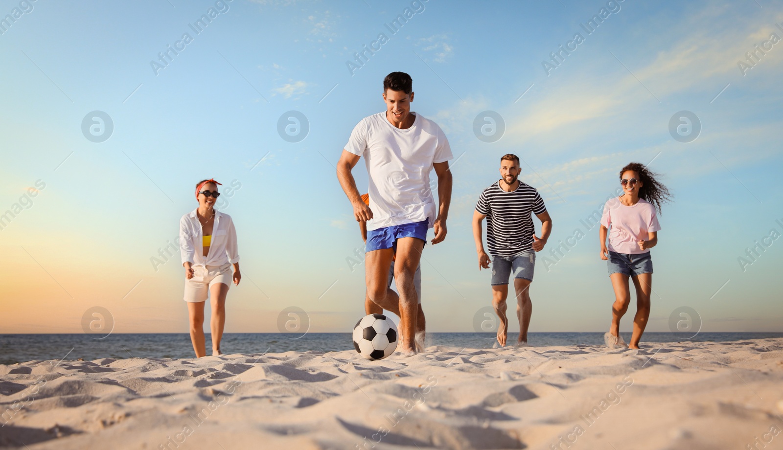 Image of Happy friends playing football on beach during sunset, low angle view