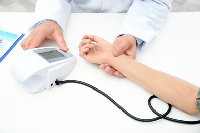 Doctor checking young woman's pulse and measuring blood pressure at table in hospital, closeup