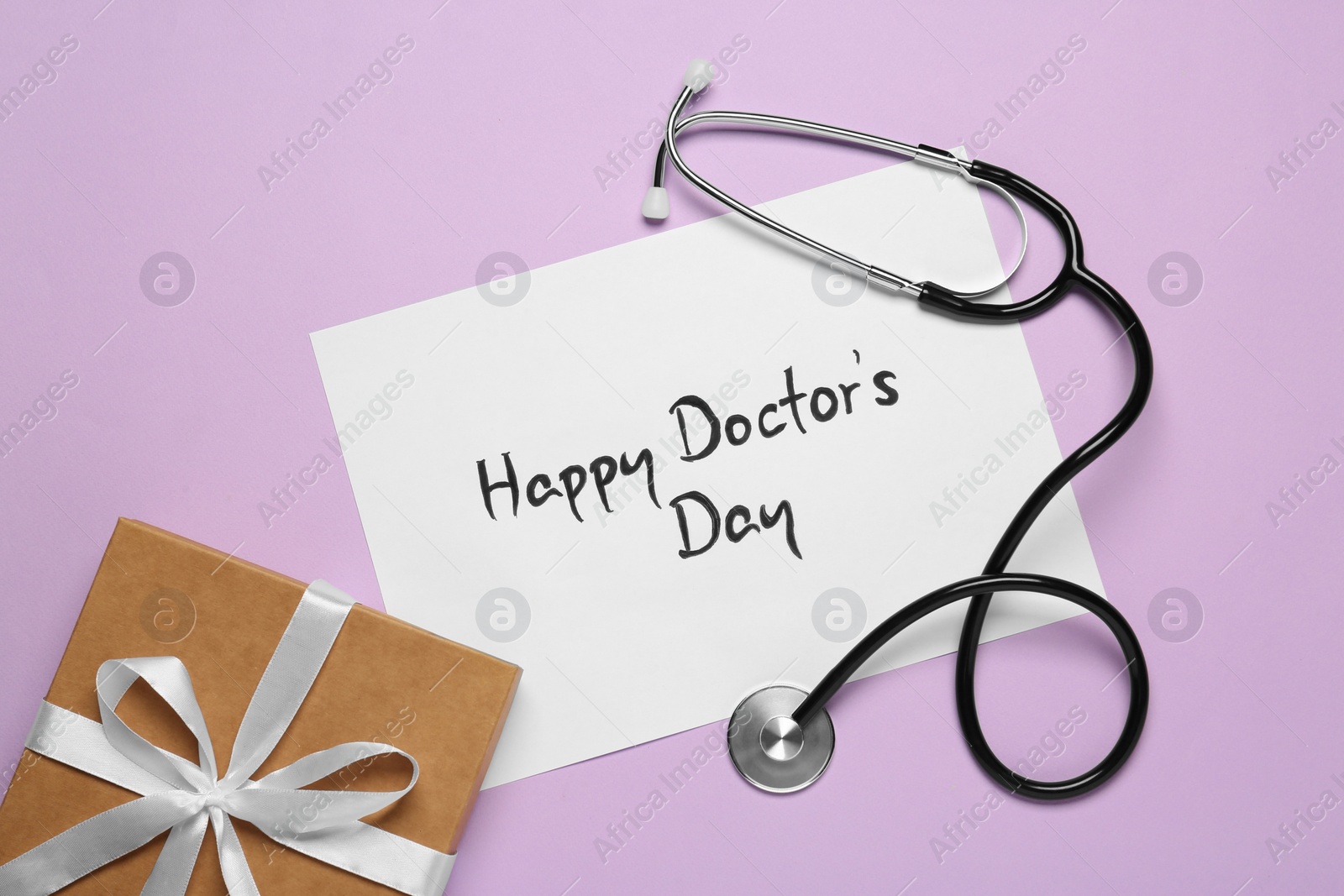 Photo of Card with phrase Happy Doctor's Day, stethoscope and gift box on light violet background, flat lay