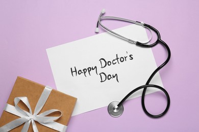 Photo of Card with phrase Happy Doctor's Day, stethoscope and gift box on light violet background, flat lay