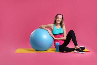 Beautiful woman sitting on yoga mat with fitness ball against pink background