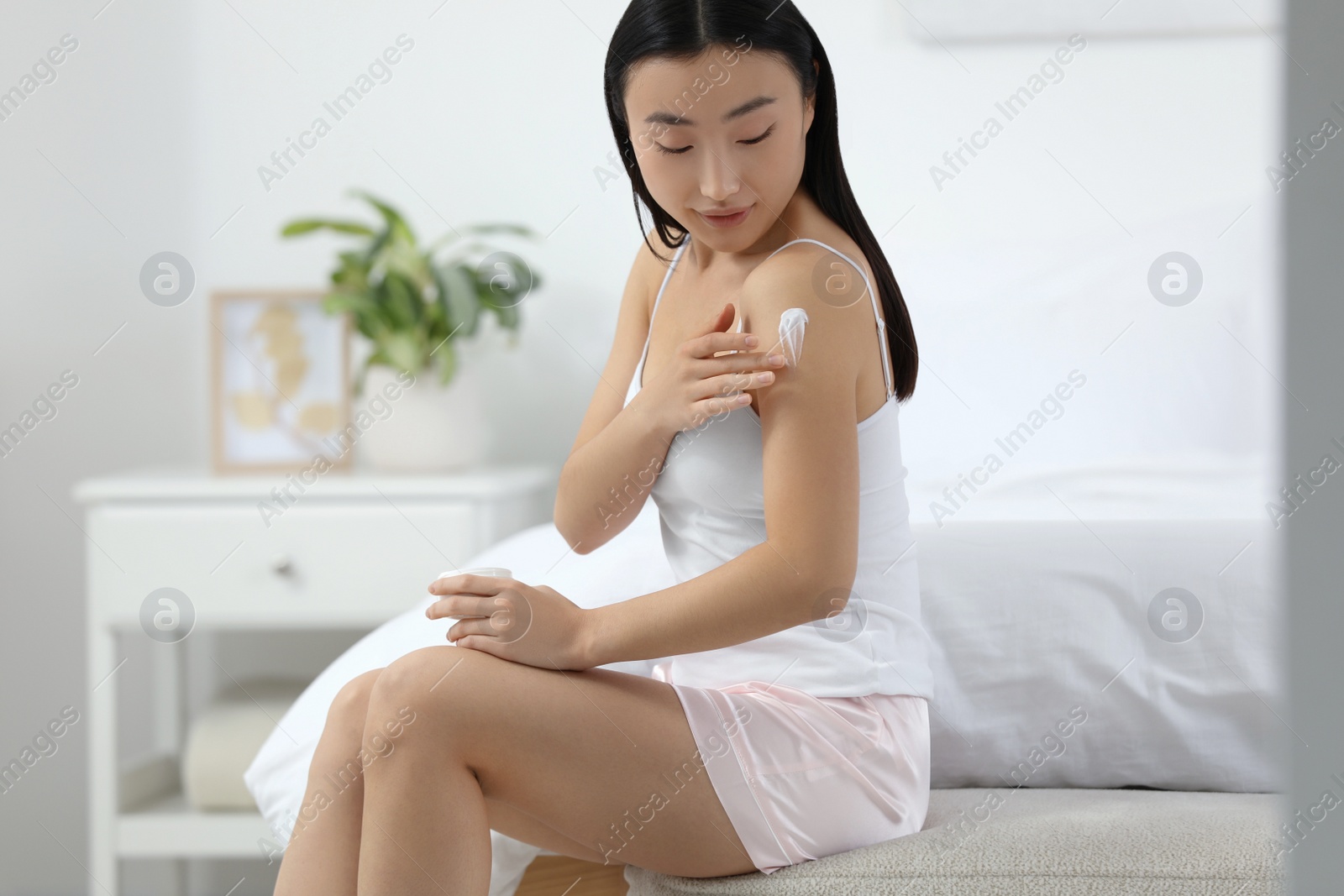 Photo of Beautiful young Asian woman applying body cream on shoulder in bedroom, space for text