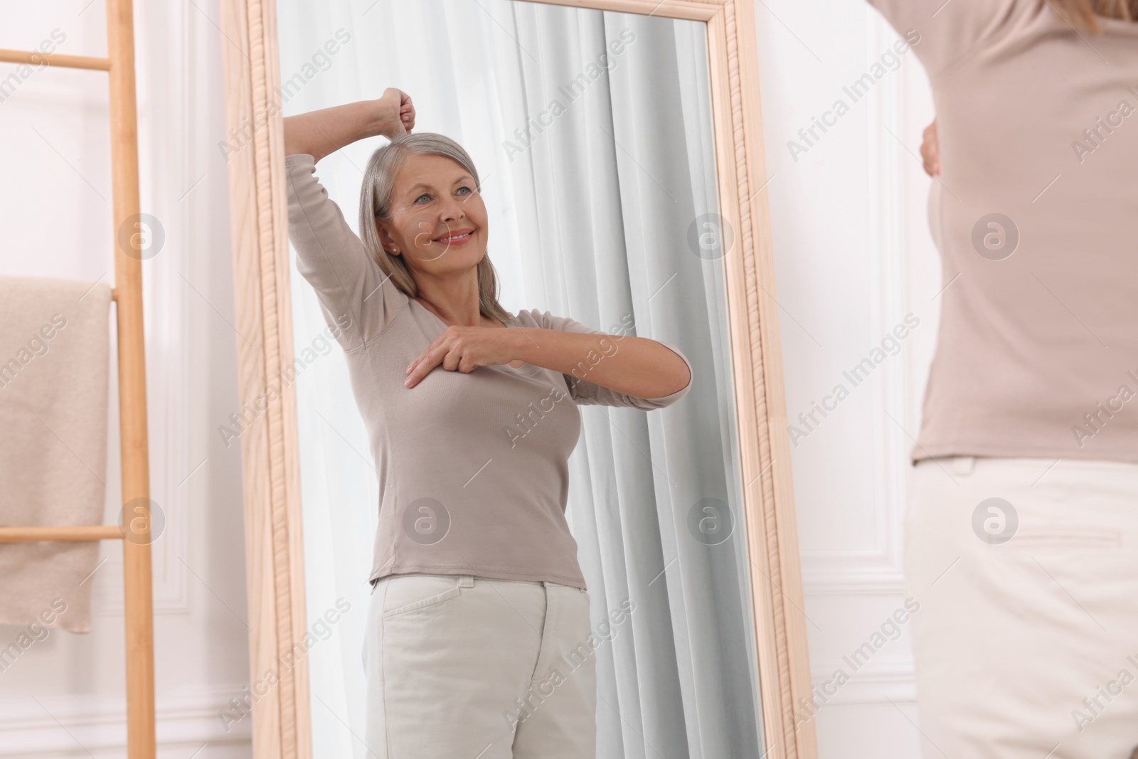 Photo of Beautiful senior woman doing breast self-examination near mirror indoors