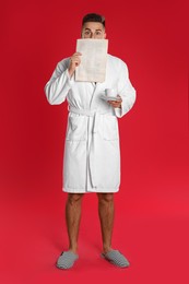 Young man in bathrobe with cup of coffee reading newspaper on red background