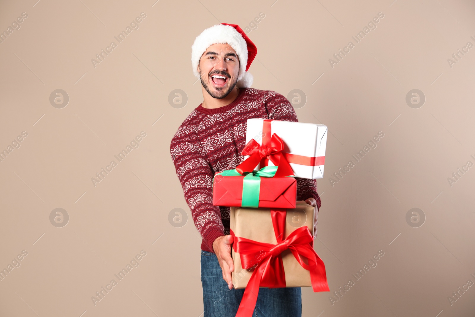 Photo of Happy man in Christmas sweater and Santa hat holding gift boxes on beige background