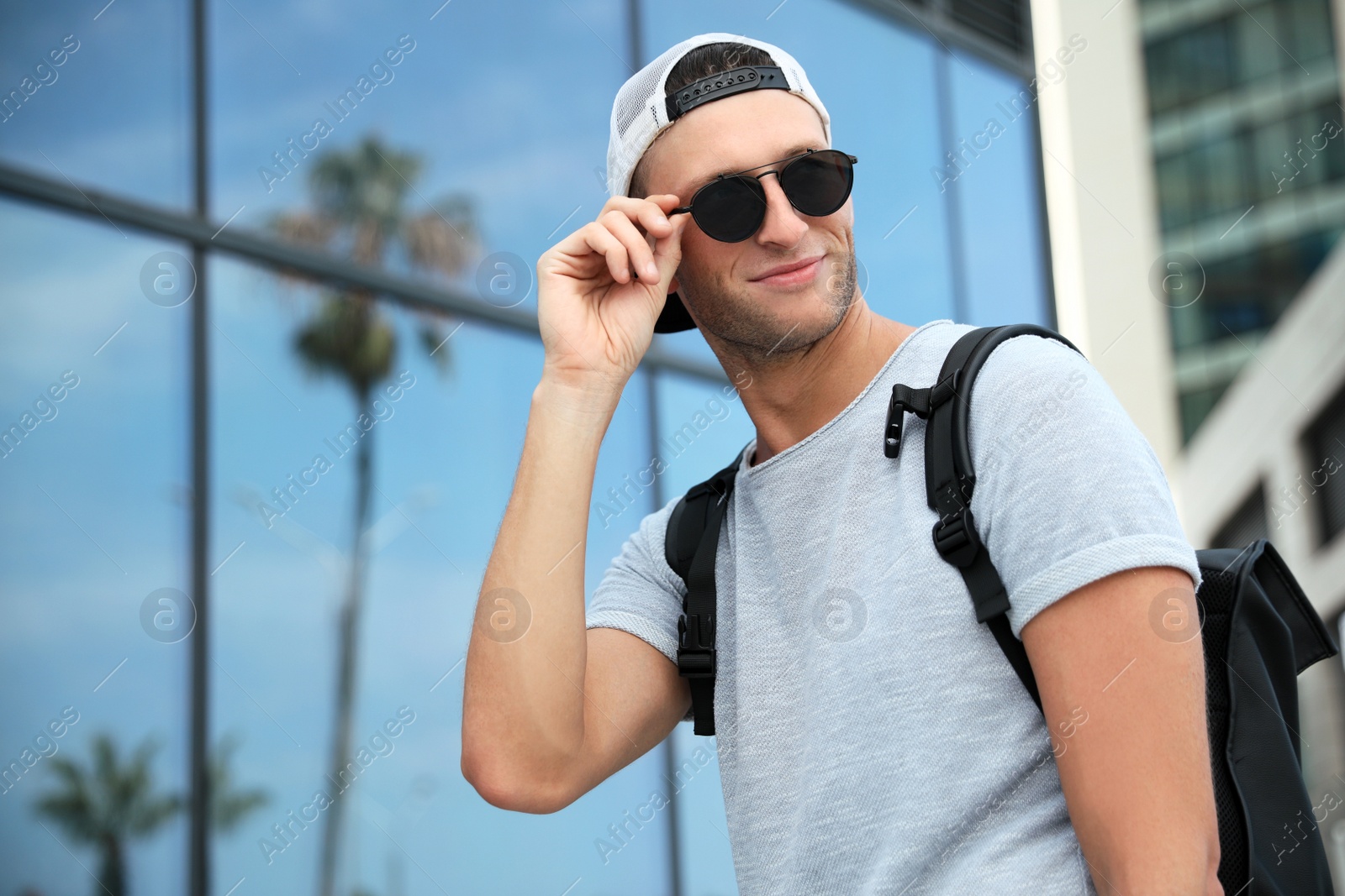 Photo of Handsome young man with stylish sunglasses and backpack near building outdoors, space for text