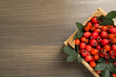 Photo of Ripe rose hip berries with green leaves in crate on wooden table, top view. Space for text