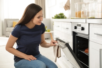 Photo of Beautiful woman opening door of oven with baked buns in kitchen