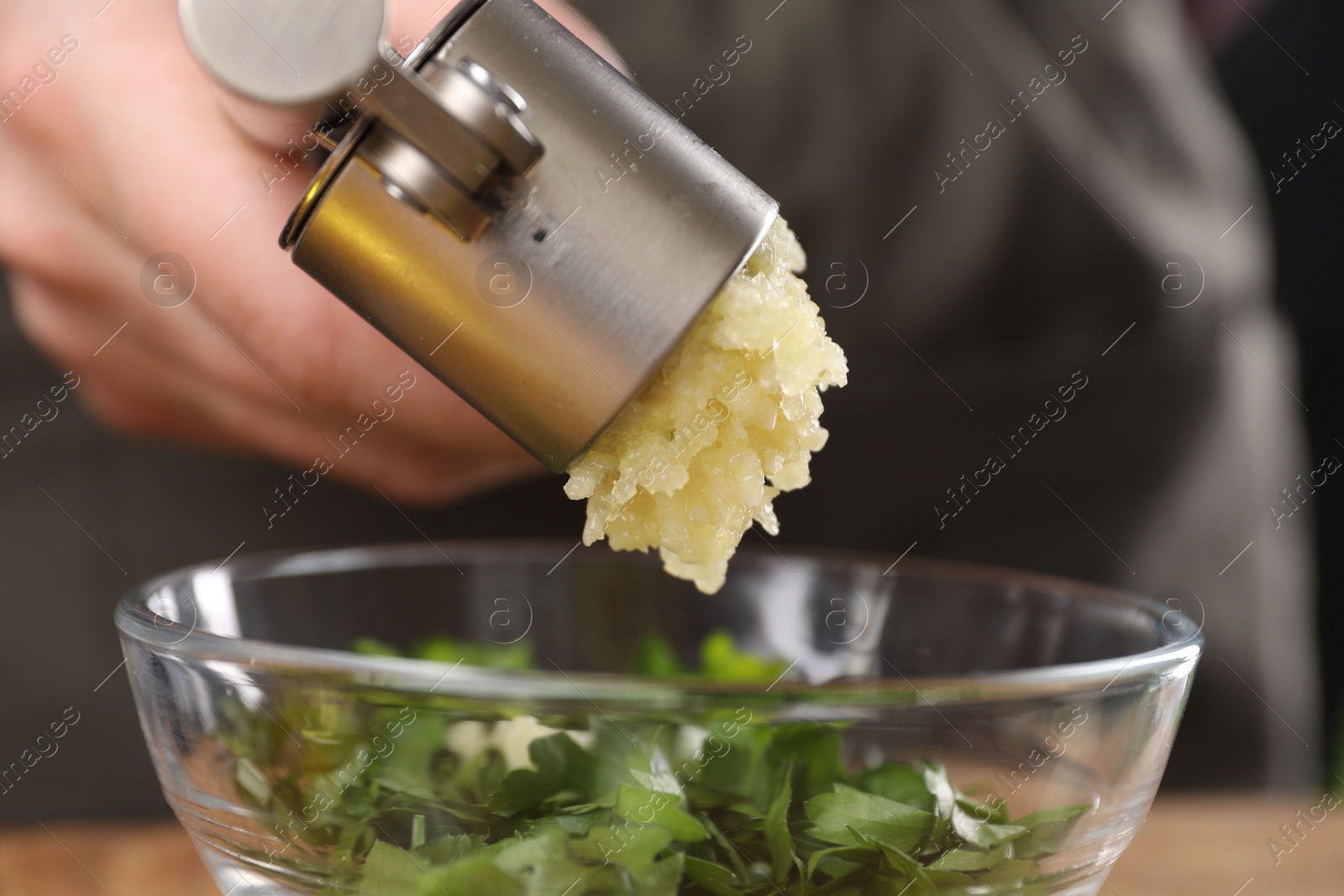 Photo of Woman squeezing garlic with press into bowl with parsley, closeup