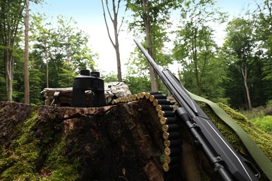 Hunting rifle, backpack, binoculars and cartridges on tree stump outdoors