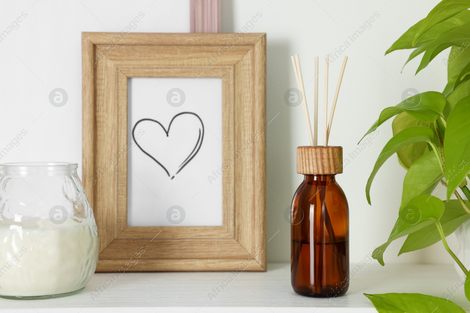 Photo of Aromatic reed air freshener, candle and wooden frame on white shelf indoors