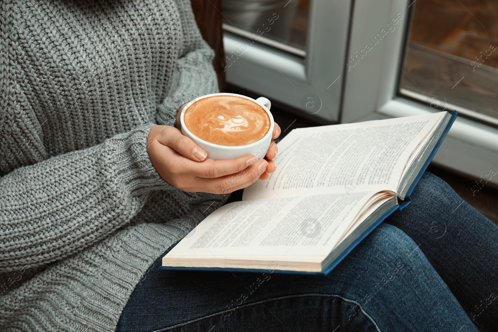 Photo of Woman with cup of coffee reading book near window indoors, closeup