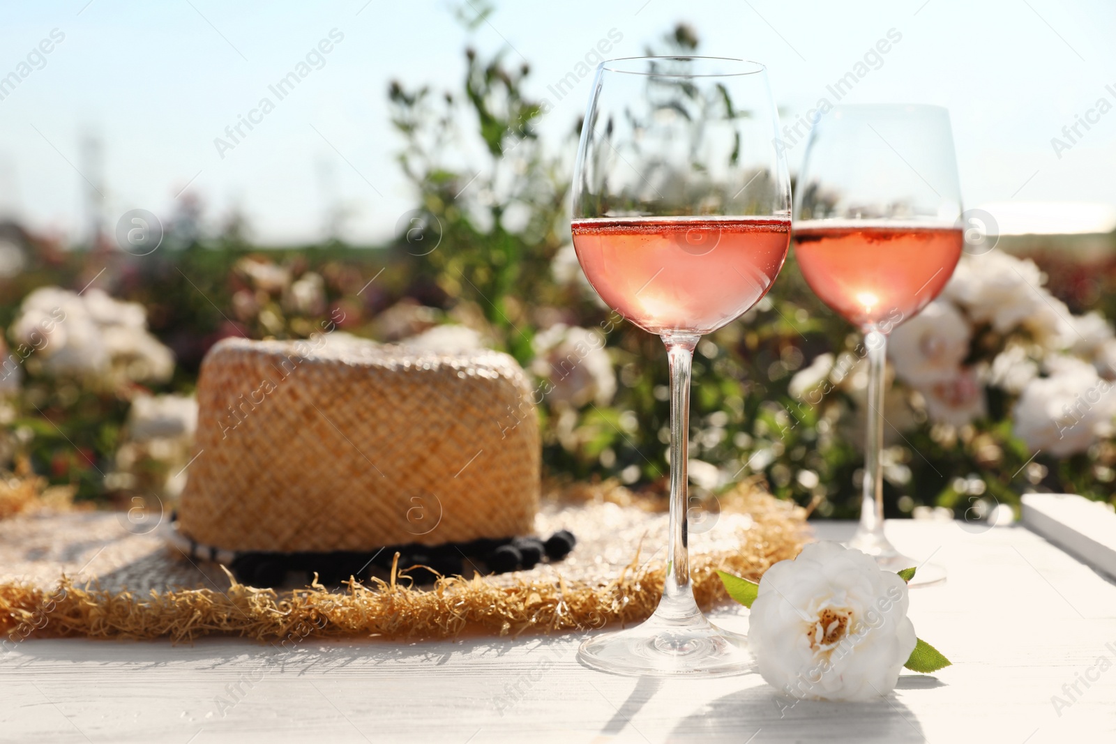 Photo of Glasses of rose wine, straw hat and beautiful flower on white wooden table outdoors. Space for text