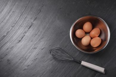Photo of Metal whisk and raw eggs in bowl on dark grey table, flat lay. Space for text