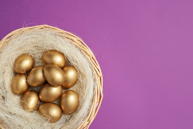 Photo of Shiny golden eggs in nest on violet background, top view. Space for text