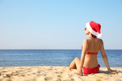 Beautiful young woman in Santa hat and bikini on beach, back view with space for text. Christmas vacation