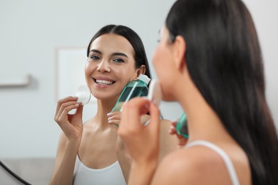 Young woman using cotton pad with micellar water near mirror indoors
