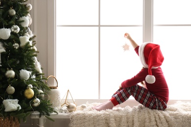 Photo of Cute little girl in Santa hat holding Christmas ornament on window sill at home