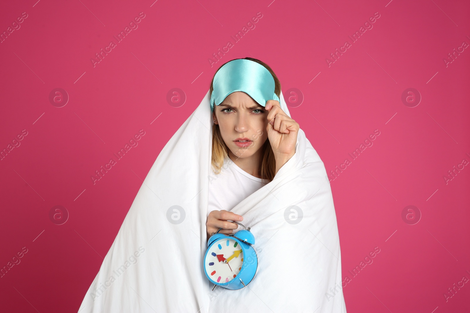 Photo of Sad young woman in sleeping mask wrapped with blanket holding alarm clock on pink background