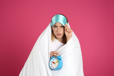 Sad young woman in sleeping mask wrapped with blanket holding alarm clock on pink background