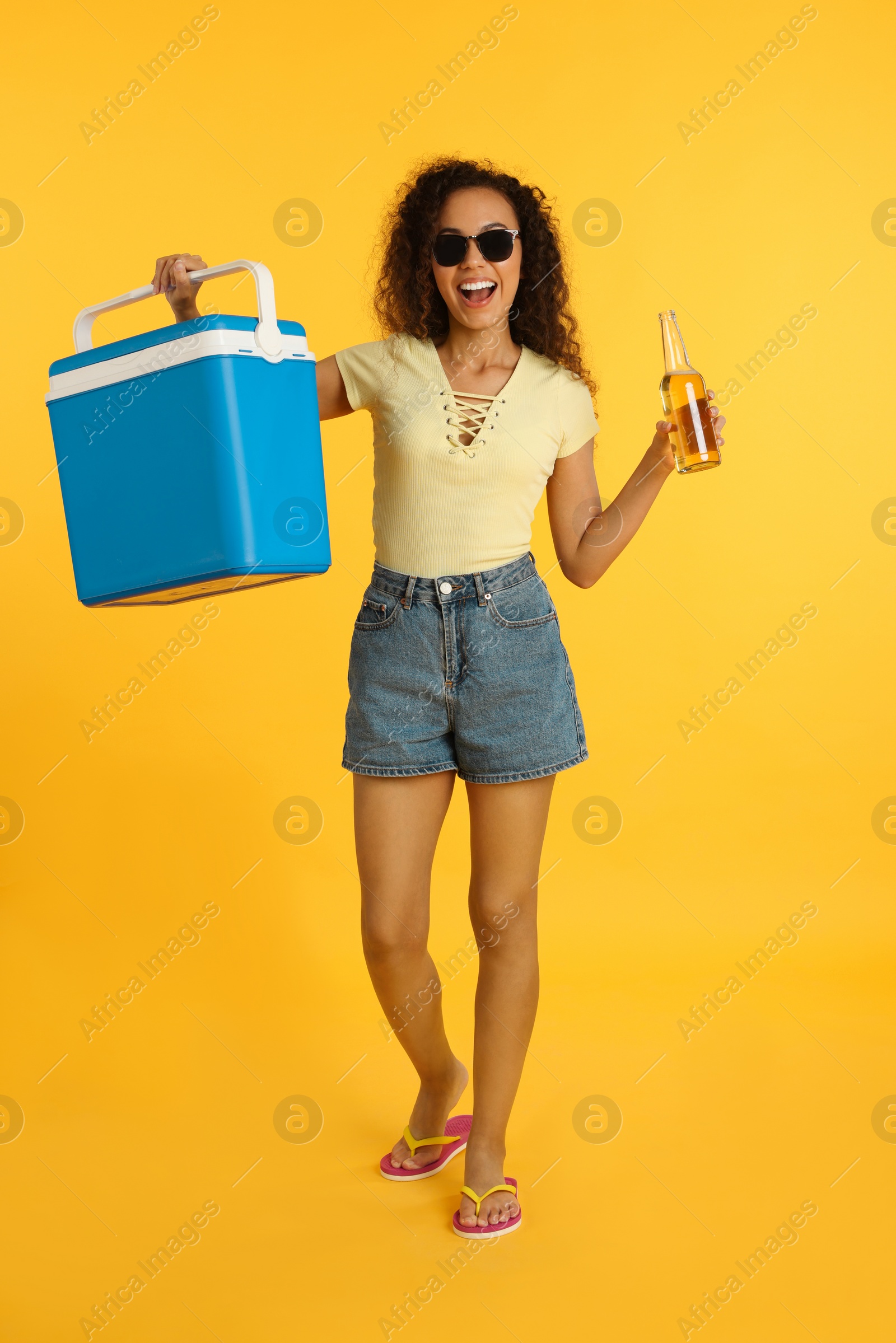 Photo of Happy young African American woman with cool box and bottle of beer on yellow background