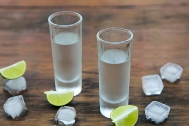 Photo of Mexican tequila shots with lime slices and ice cubes on wooden table, closeup. Drink made from agave