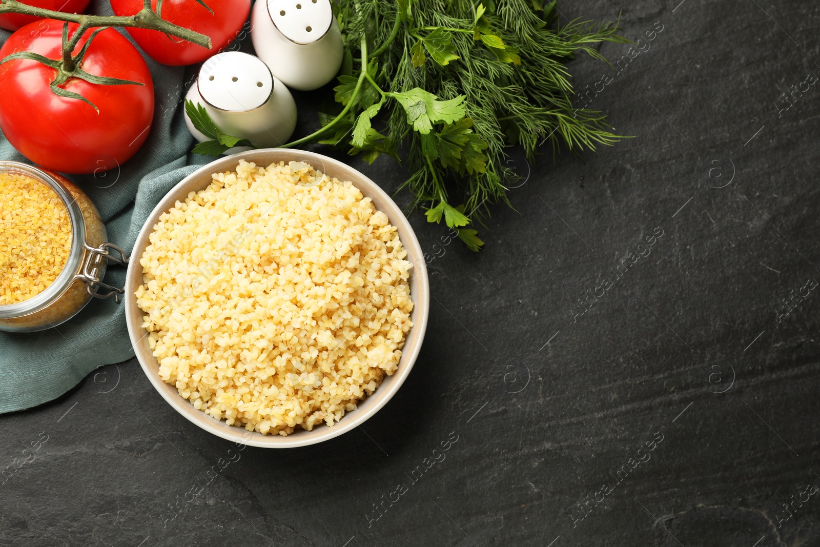 Photo of Delicious bulgur in bowl, dill, parsley and tomatoes on black table, top view. Space for text