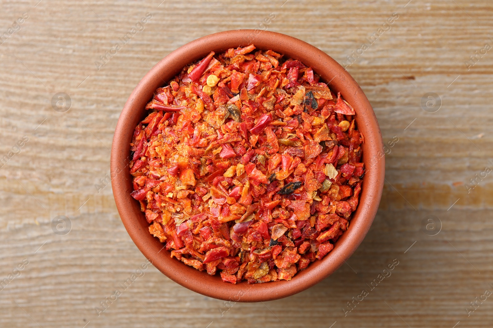 Photo of Aromatic spice. Red chili pepper flakes in bowl on wooden table, top view