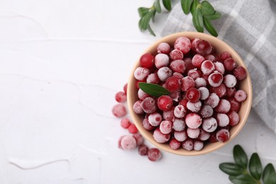 Photo of Frozen red cranberries in bowl and green leaves on white table, top view. Space for text