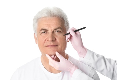 Doctor drawing marks on mature man's face for cosmetic surgery operation against white background