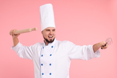 Photo of Happy professional confectioner in uniform holding rolling pin and whisk on pink background