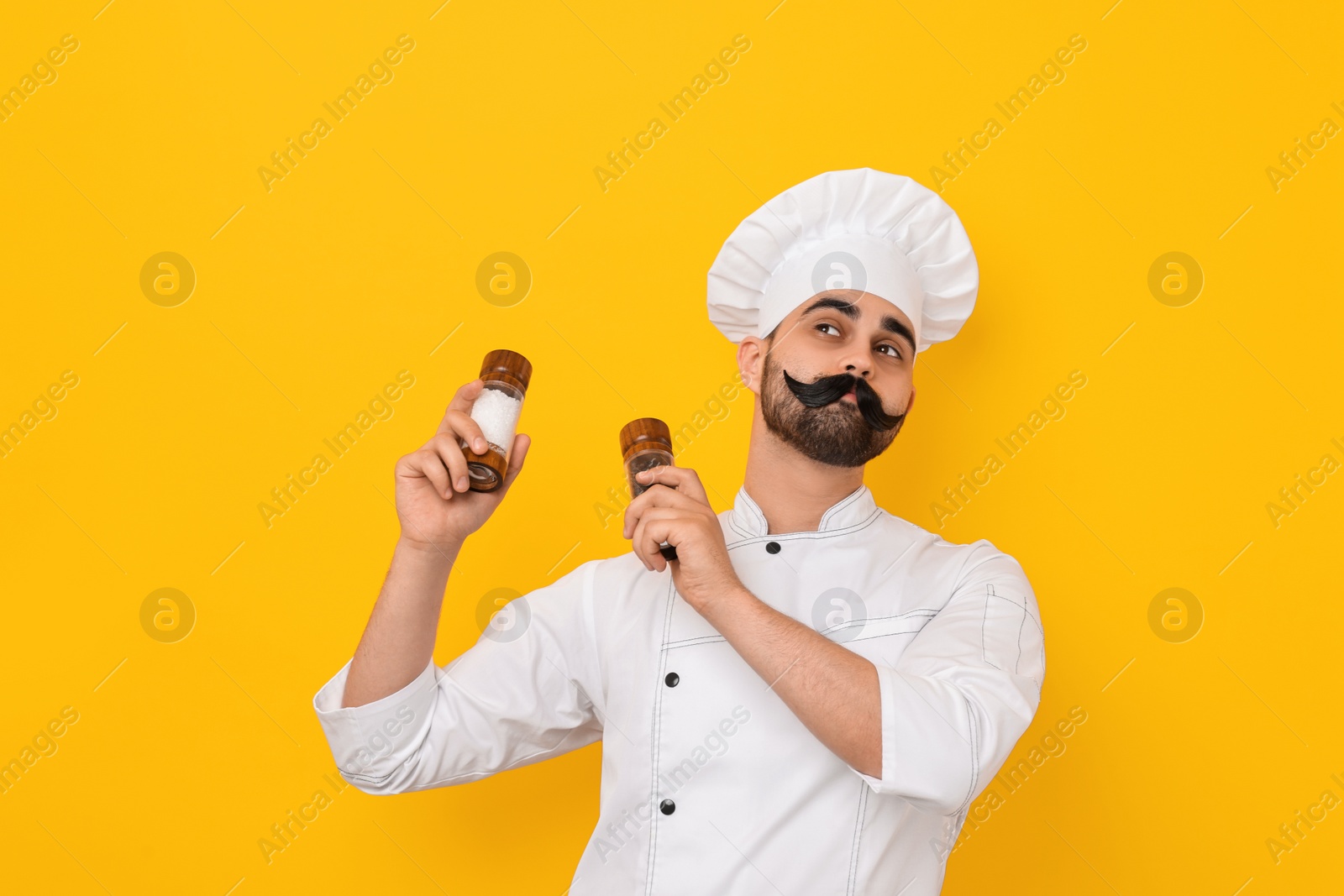 Photo of Professional chef with funny artificial moustache holding shakers on yellow background