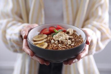 Woman holding bowl of tasty granola with banana, cashew and strawberries indoors, closeup