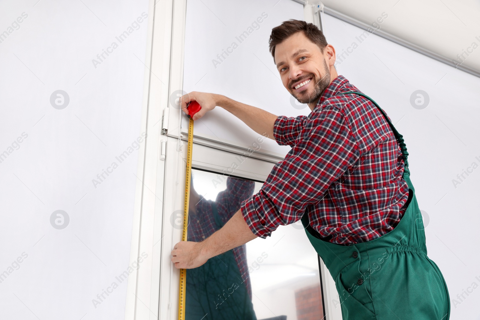 Photo of Worker in uniform using tape measure while installing roller window blind indoors