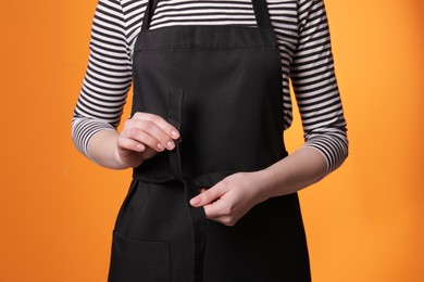 Photo of Woman putting on black apron against orange background, closeup