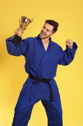 Photo of Portrait of happy young man in blue kimono with gold trophy cup on yellow background