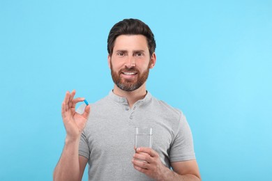 Handsome man with glass of water and pill on light blue background