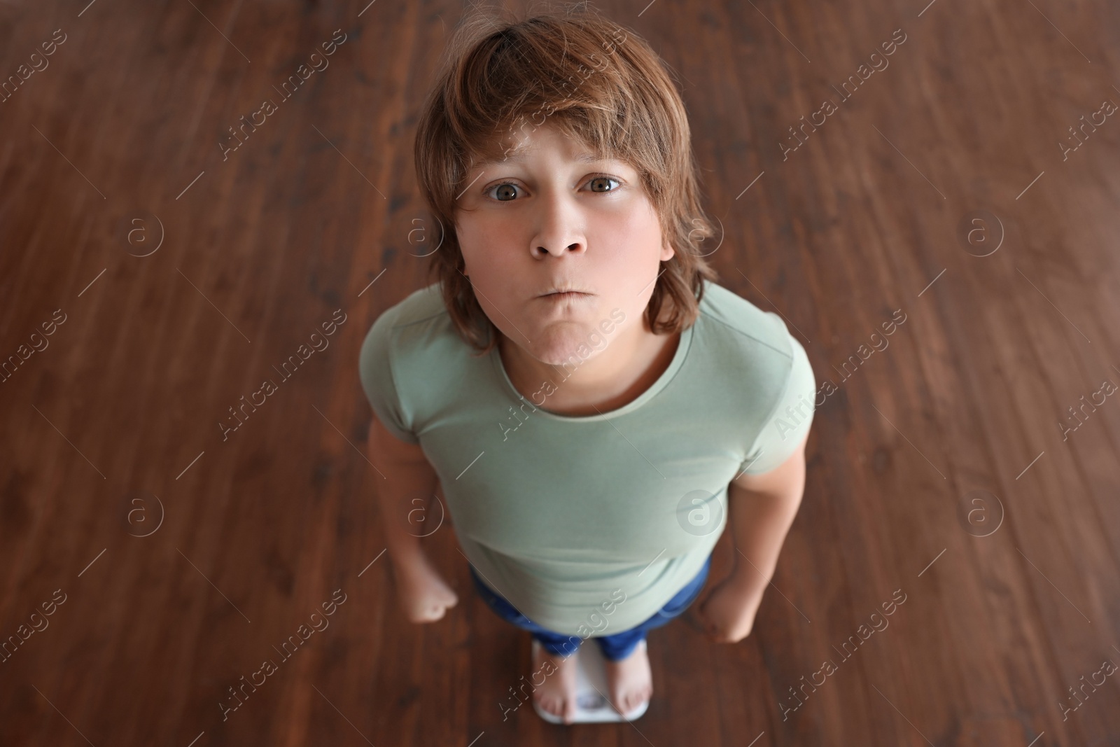 Photo of Emotional overweight boy standing on floor scales indoors, above view