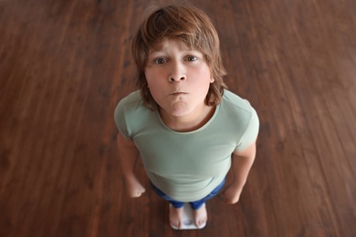 Photo of Emotional overweight boy standing on floor scales indoors, above view