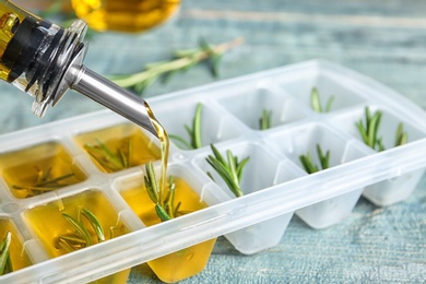 Photo of Pouring olive oil into ice cube tray with rosemary on table, closeup